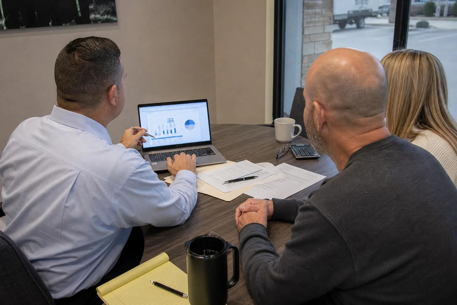 Agent in a light blue button-down shirt reviews charts on a laptop with a couple at a round conference table beside a storefront window