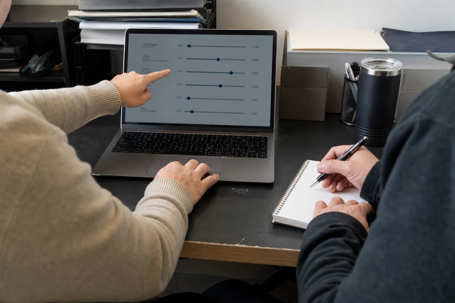 Two people at an office desk: one points to slider options on a laptop screen while the other writes notes in a spiral notebook