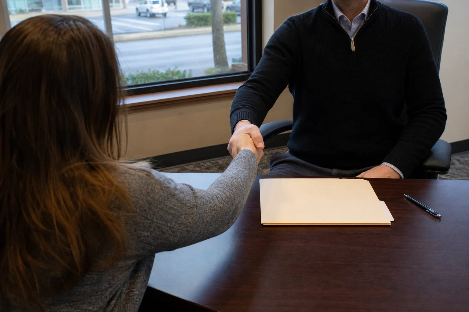 An agent in a dark sweater shakes hands with a client across a wooden desk holding a closed manila folder and pen, with a window overlooking a parking lot.